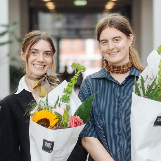 De to smilende prisvindere Helene Franch og Feline Kamber holder blomsterbuketter foran rækker af stole.